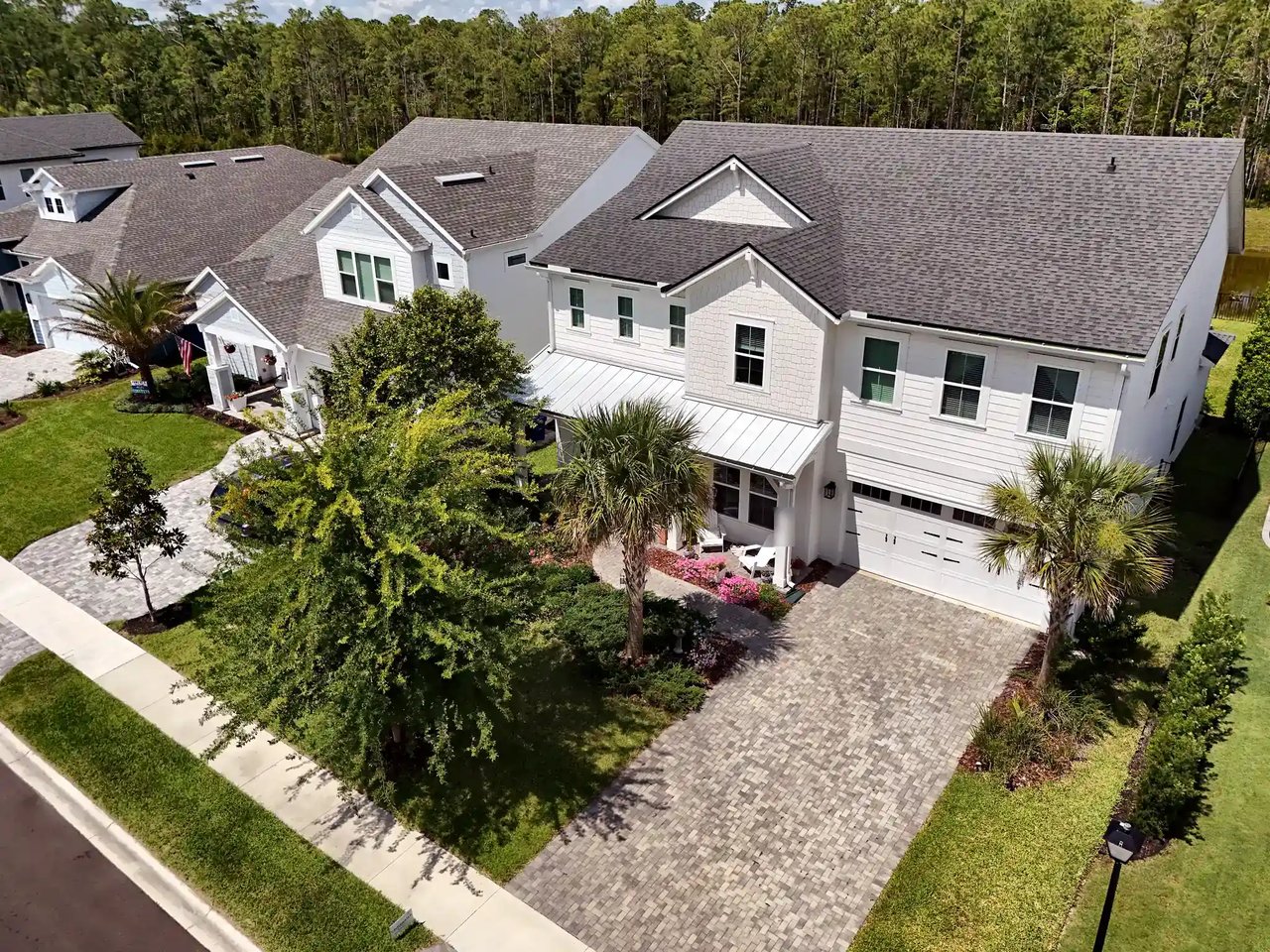 Elevated view of a white two-story coastal-style home with a metal-roof porch, palms and a paver driveway in Palm Crest.