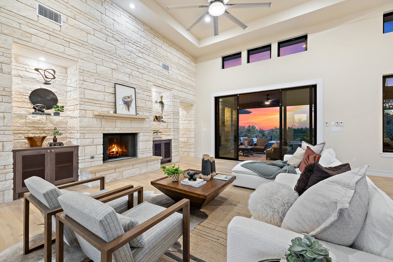Living room with a stone fireplace and sliding glass doors opening to a sunset view, in this Bee Caves home on Caudill Lane, Austin, Texas.