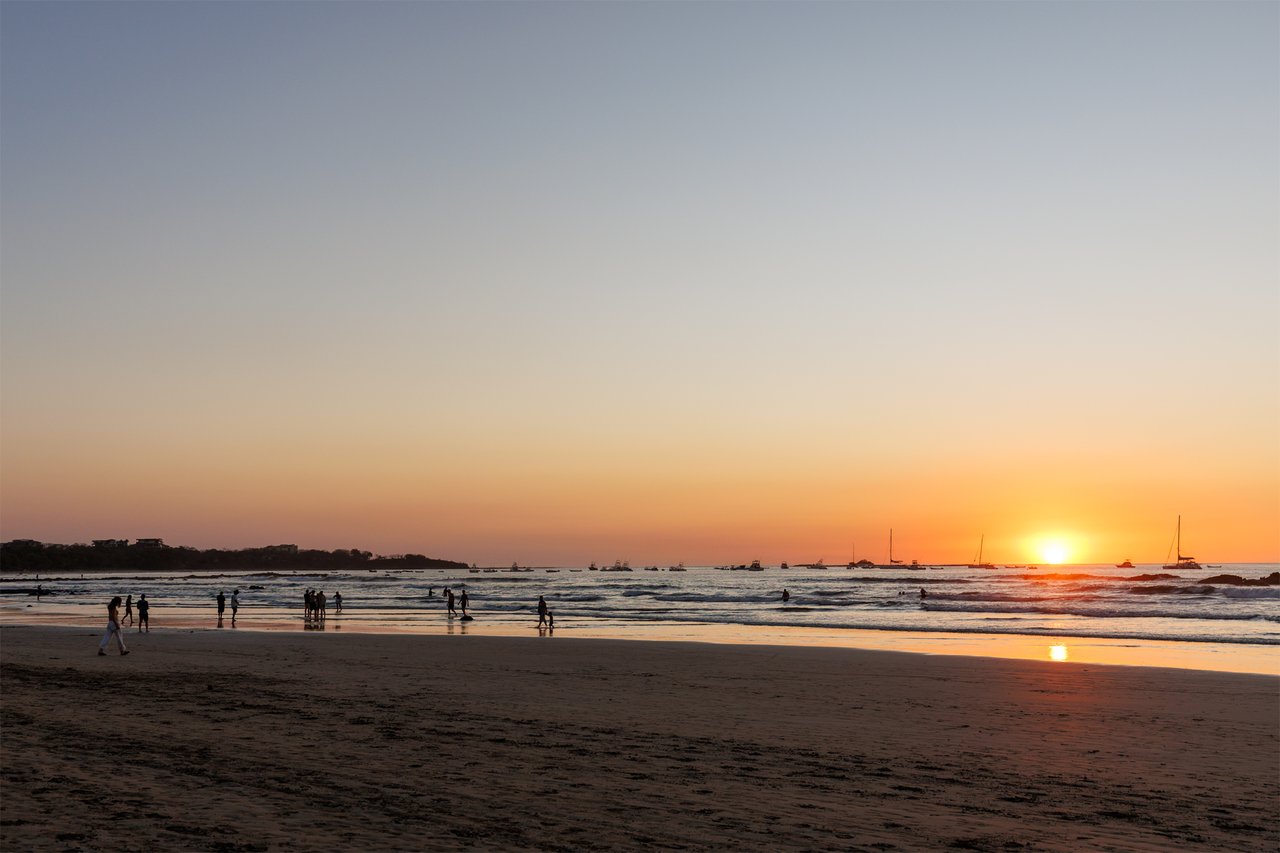Beachgoers stroll along the shore as boats sit offshore beneath a glowing sunset.