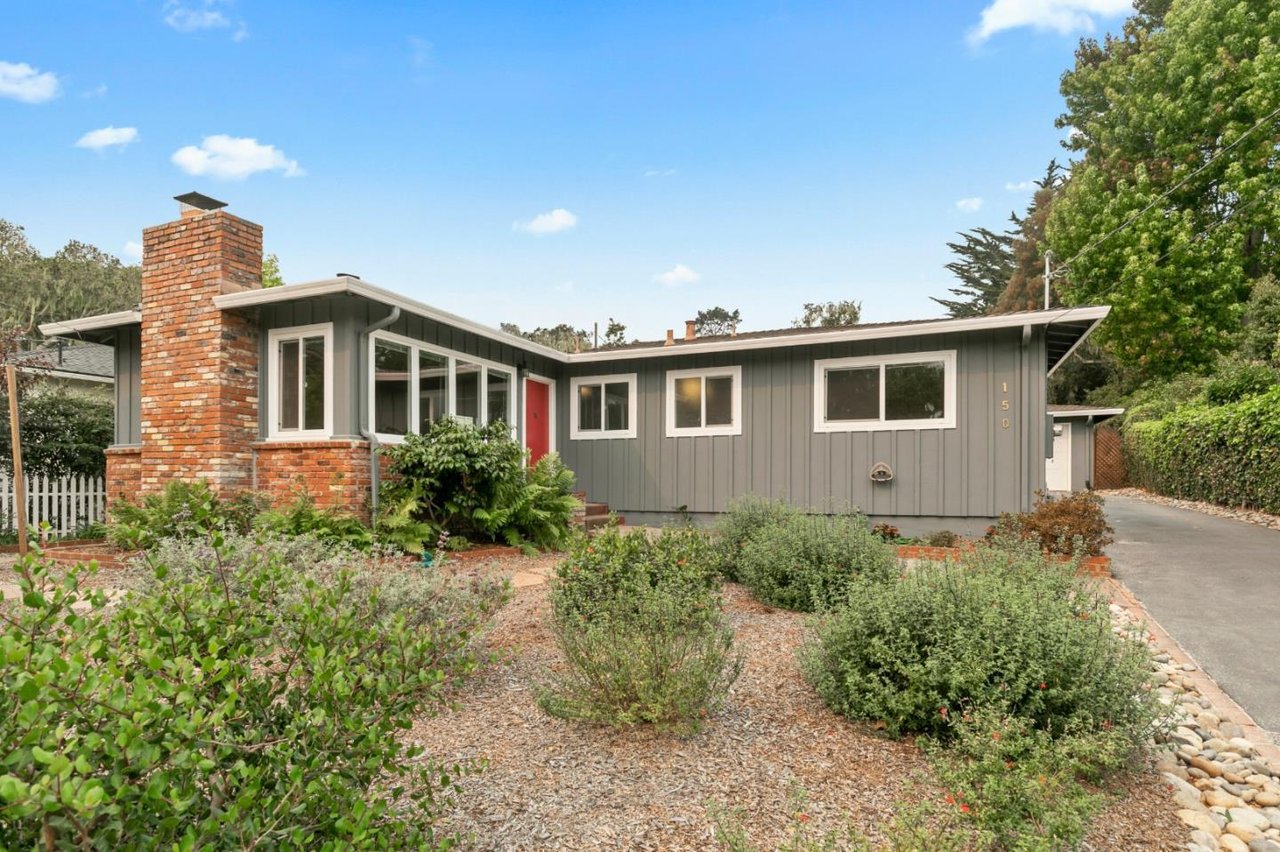 Gray single-story home with a brick chimney, red front door, and landscaped garden.