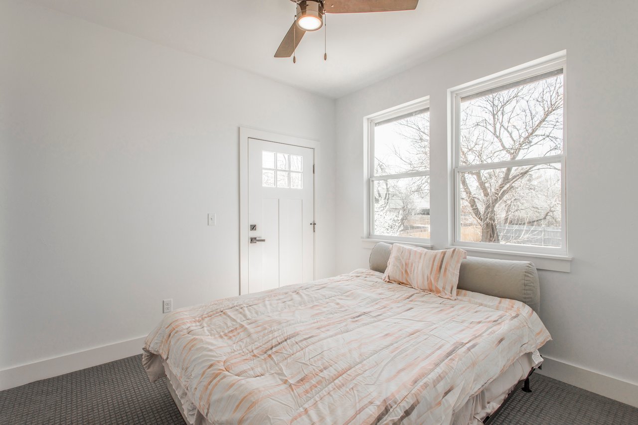 Furnished bedroom with pale bedding, two large windows overlooking bare trees, and a ceiling fan.