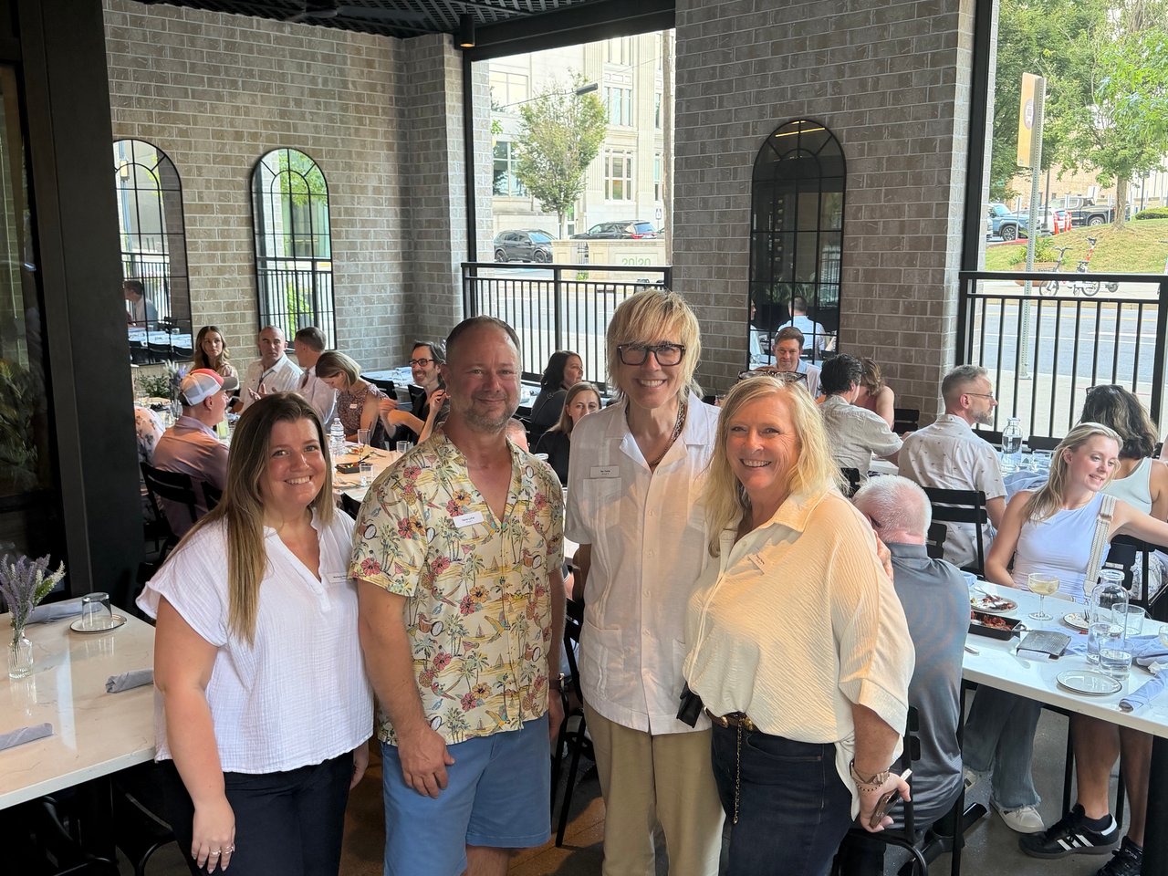 Four people pose together at a restaurant gathering, with diners and arched windows behind them.