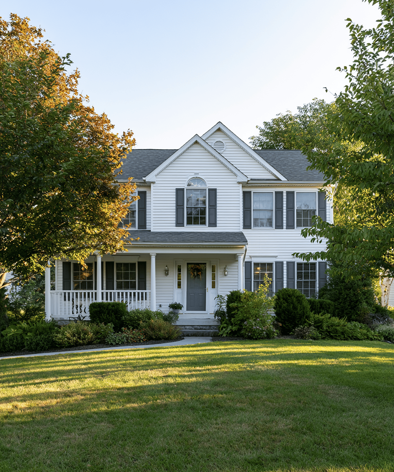 White two-story house with a covered porch, dark shutters, and autumn trees around the lawn.