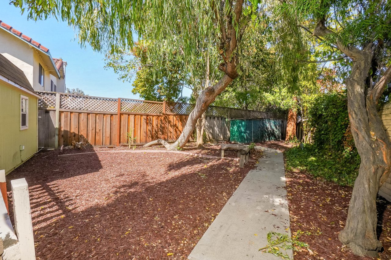 Backyard with mature shade trees, mulched ground, a concrete path, and a wooden privacy fence.