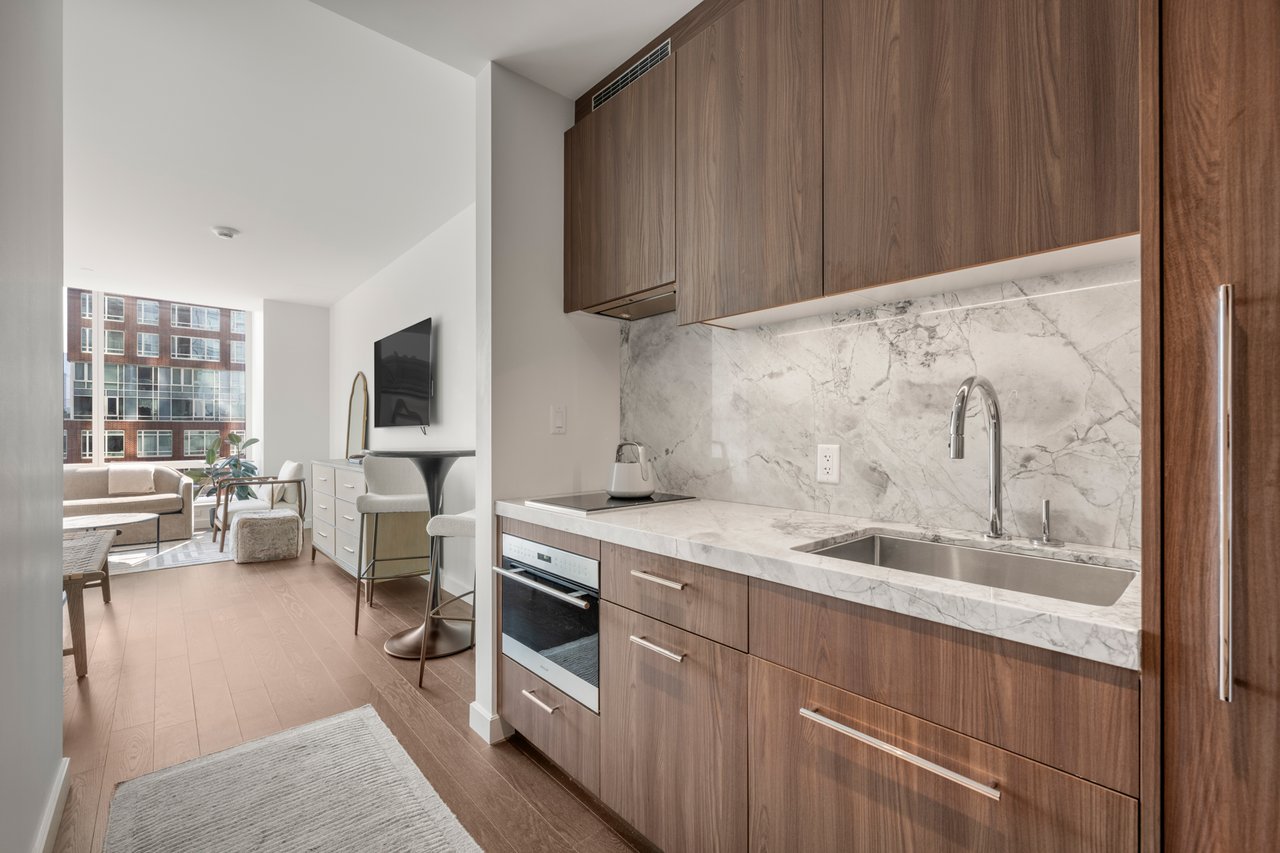 Modern apartment kitchen with walnut cabinets, veined stone counters, and a living area beside large windows.