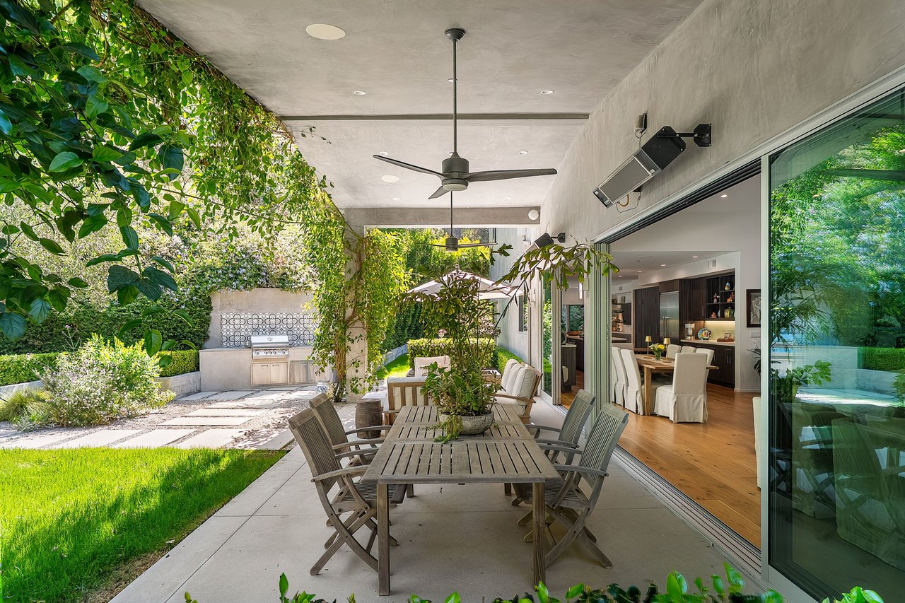 Covered patio with a long outdoor dining table, built-in grill, sliding glass doors, and lush garden.