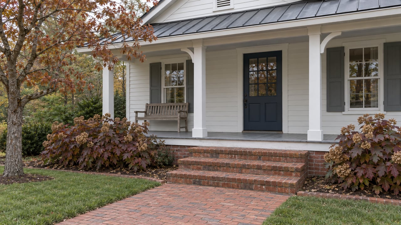 Brick approach and steps leading to a white cottage porch with a dark blue door, weathered metal roof and autumn dogwood foliage.