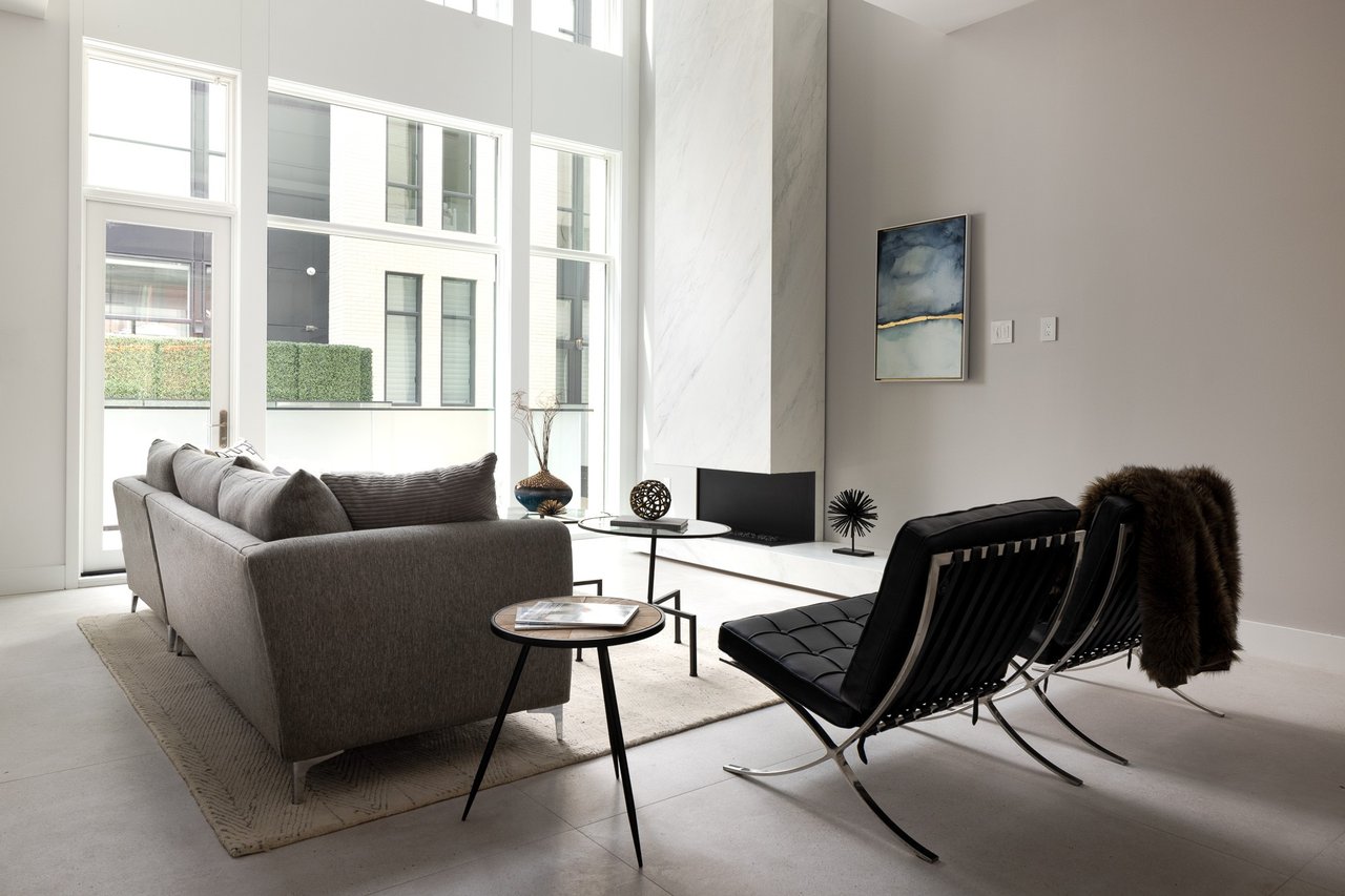 Furnished living room with a gray sofa, black lounge chairs, and a tall marble fireplace wall.