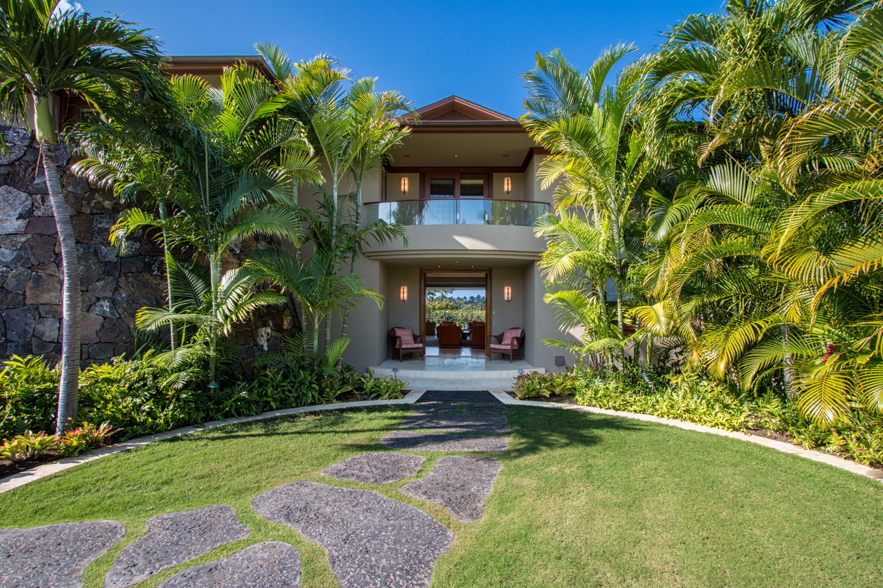 Two-story tropical home framed by palms, with a glass balcony and stone path.
