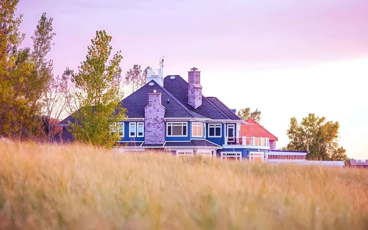 Blue-sided house with stone chimneys beyond a golden field at sunset.