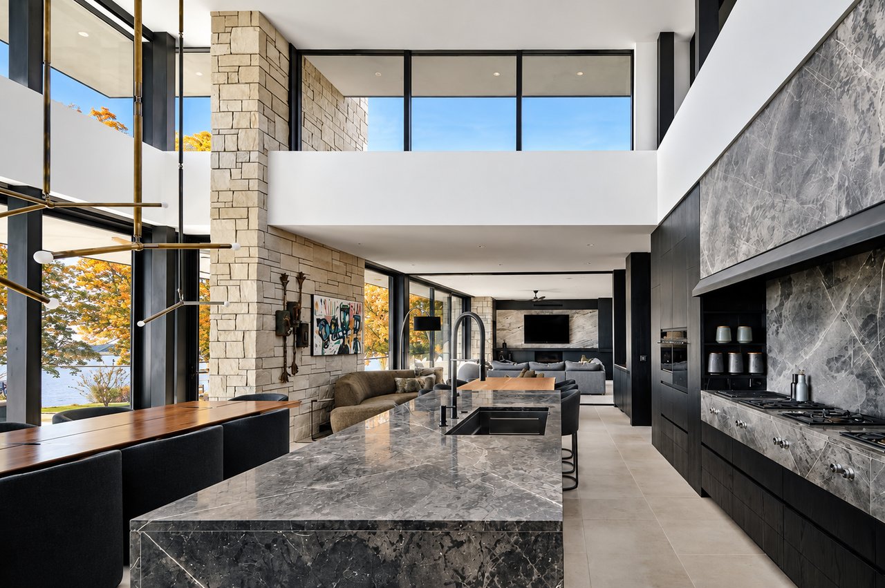 Modern kitchen with a marble island and expansive windows overlooking water and autumn foliage.