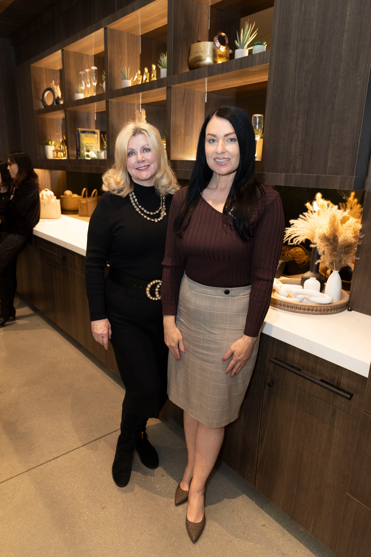 Two women pose together at an open house beside dark wood display shelves; a third woman sits in the background.