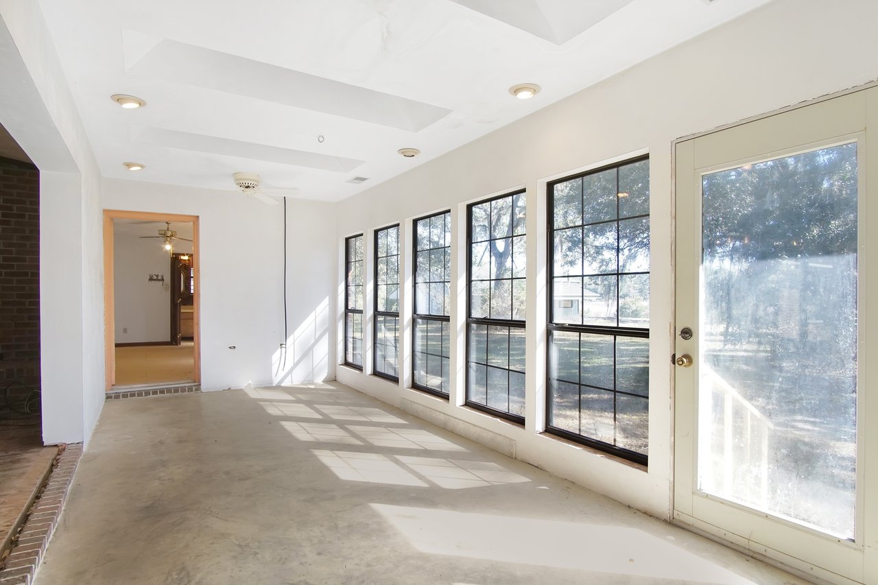 Bright sunroom with large windows and glass door on the right, casting shadows on the concrete floor. Ceiling features recessed lights and a fan. Mood is serene.