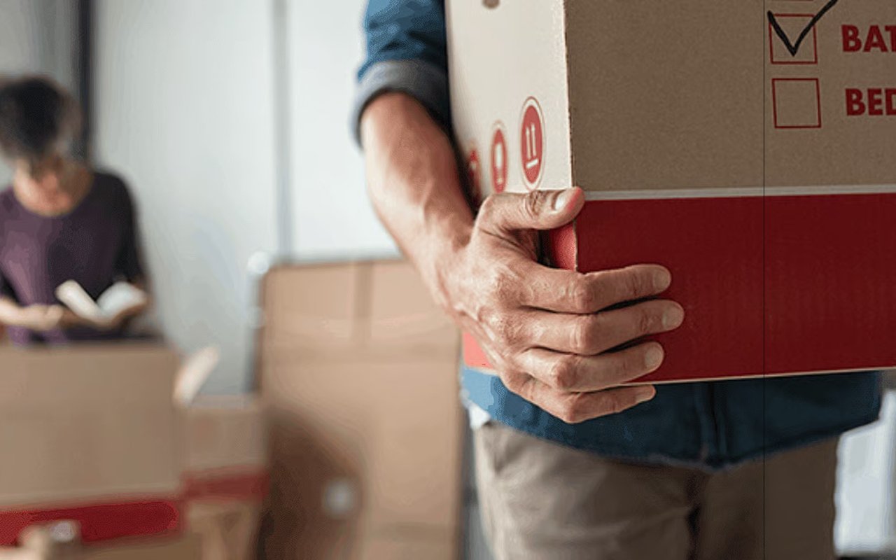 A person carries a large moving box as another person packs boxes in the background.