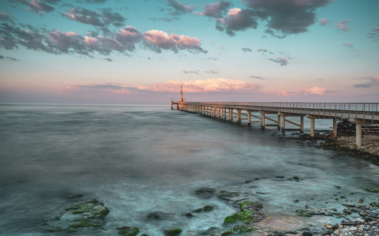 Concrete pier stretching over calm water beside a rocky shoreline at sunset.