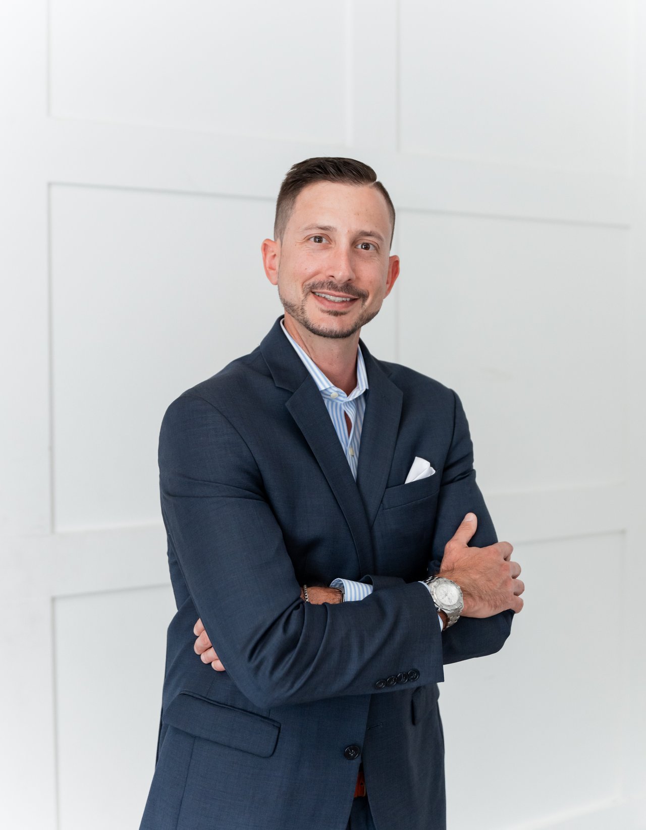 Bryan Waggoner in a navy suit, standing with his arms crossed against a light paneled wall.