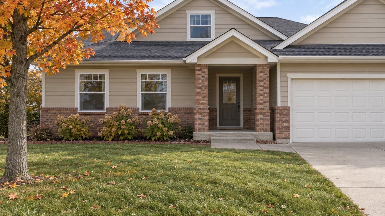 Autumn lawn and concrete walkway leading to a tan-sided home with brick porch columns and orange maple foliage.