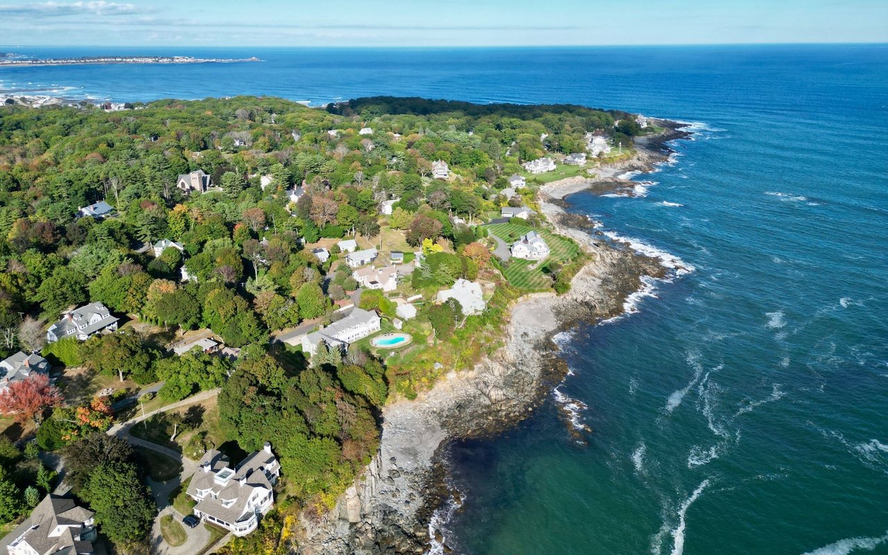 Aerial view of homes among trees along a rocky coastline, with a turquoise pool.