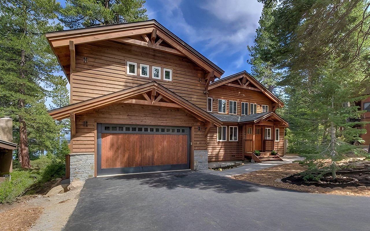 Two-story wood-sided mountain home with a wide garage door and paved driveway among evergreens.