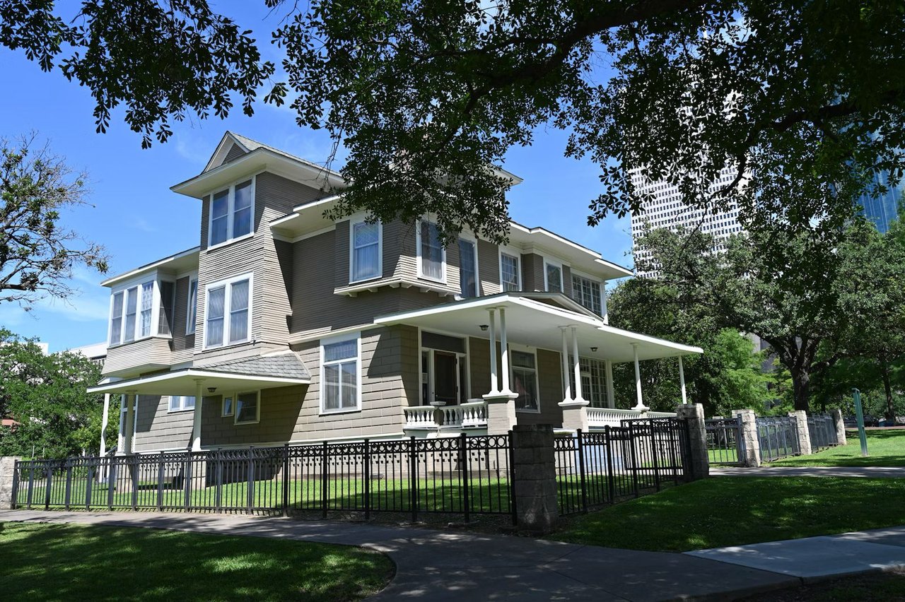 Two-story gray house with a covered porch, black metal fence, and mature trees.