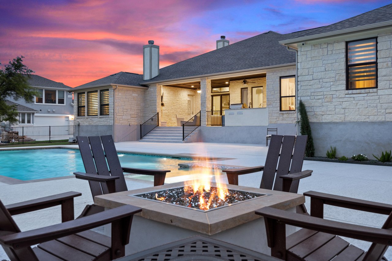 Twilight exterior of a luxury stone home in Grand Mesa, Leander, Texas, featuring a backyard pool, covered patio, and fire pit seating area.