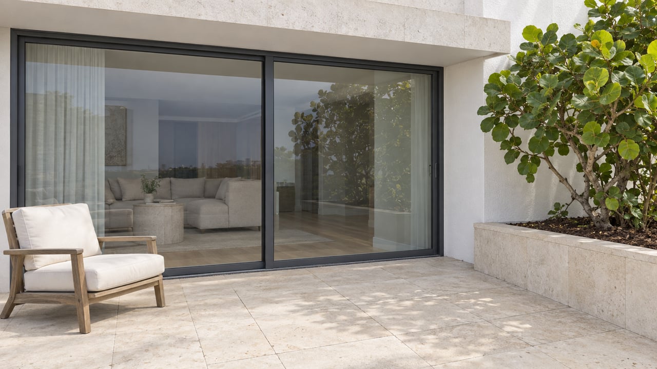 A private apartment terrace with cream limestone paving, a teak chair, closed glass doors and a sea-grape tree in a raised stone planter.
