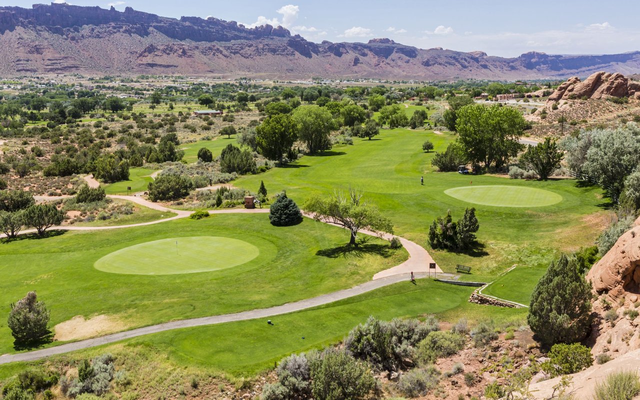 Golf course with bright green putting greens and paths beneath a rugged mountain range.