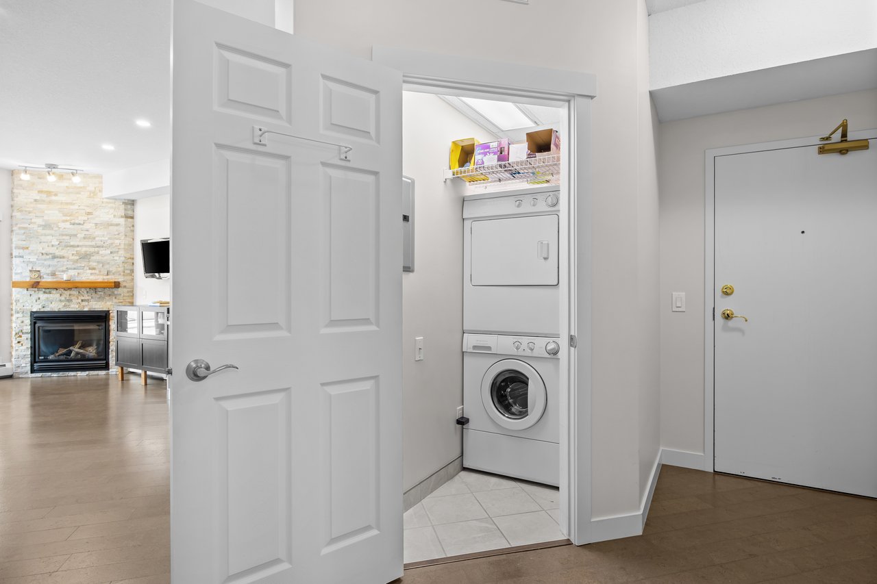 Stacked washer and dryer in a laundry closet beside the condo entry; a stone fireplace is visible beyond.