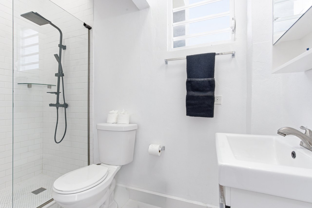 White bathroom with a glass shower, black fixtures, toilet, and sink beneath a window.