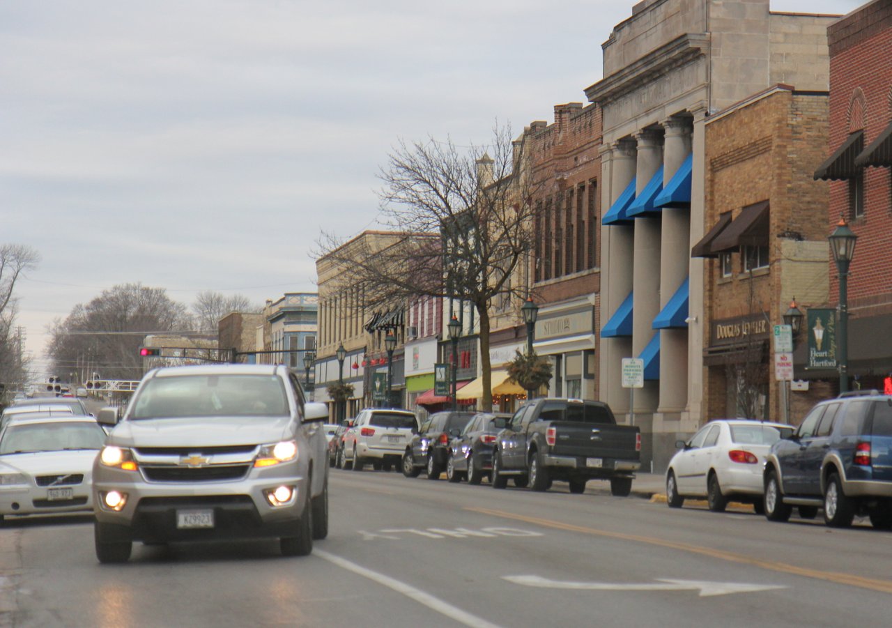 Cars driving through downtown Hartford, Wisconsin with storefronts and buildings along Main Street.