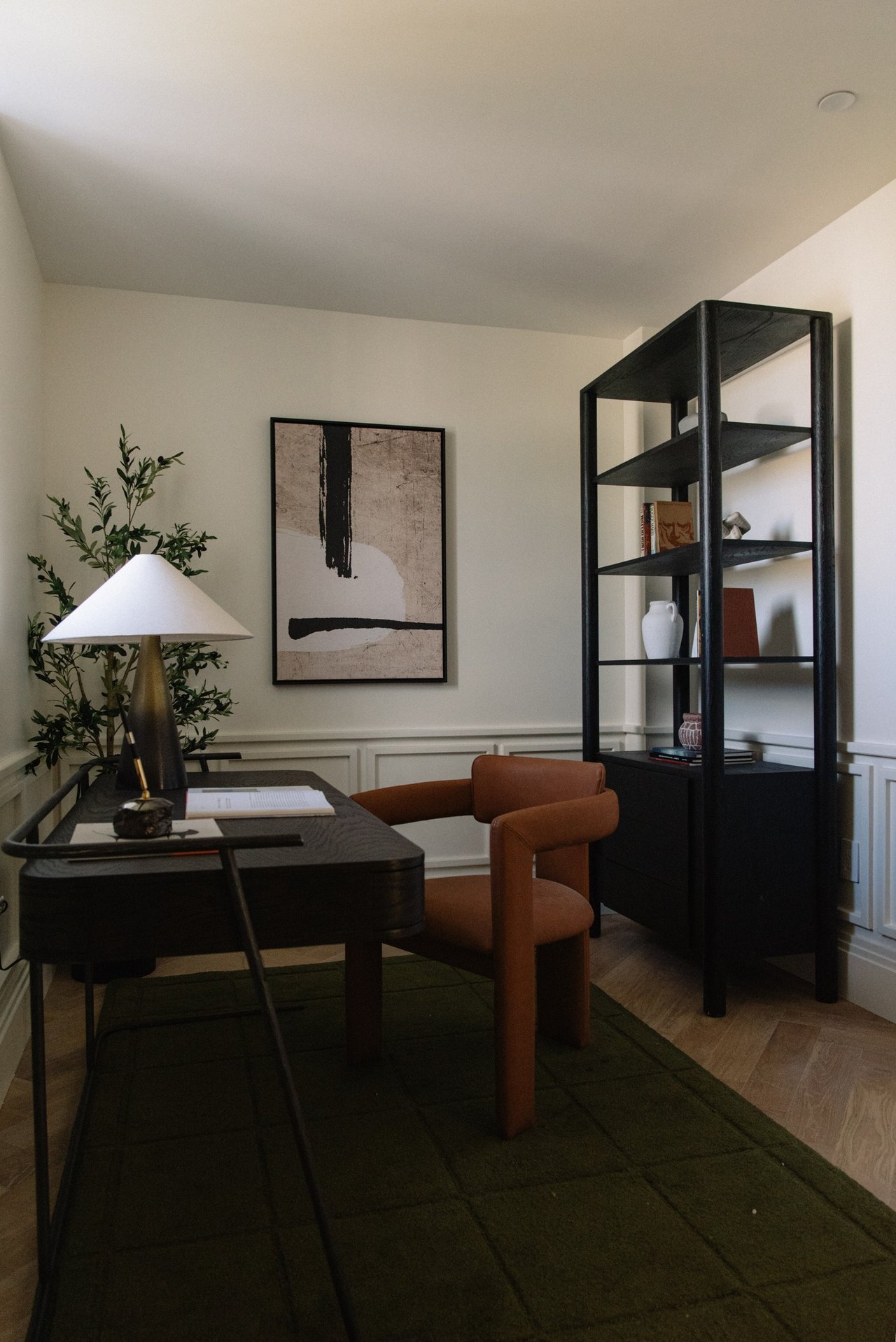 Furnished office with a dark desk, rust-colored chair, black shelving, and framed abstract art.