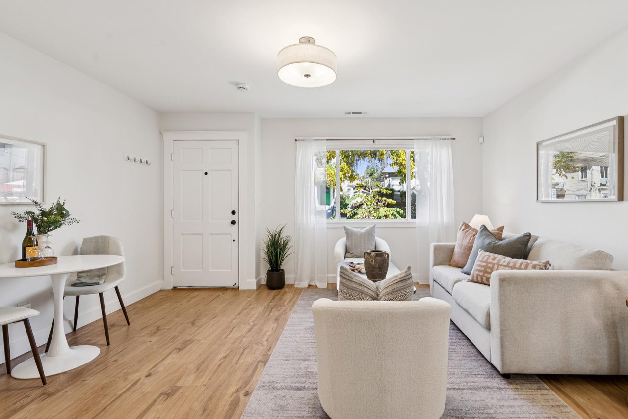 Staged living room with light wood floors, white seating, and a window overlooking greenery.