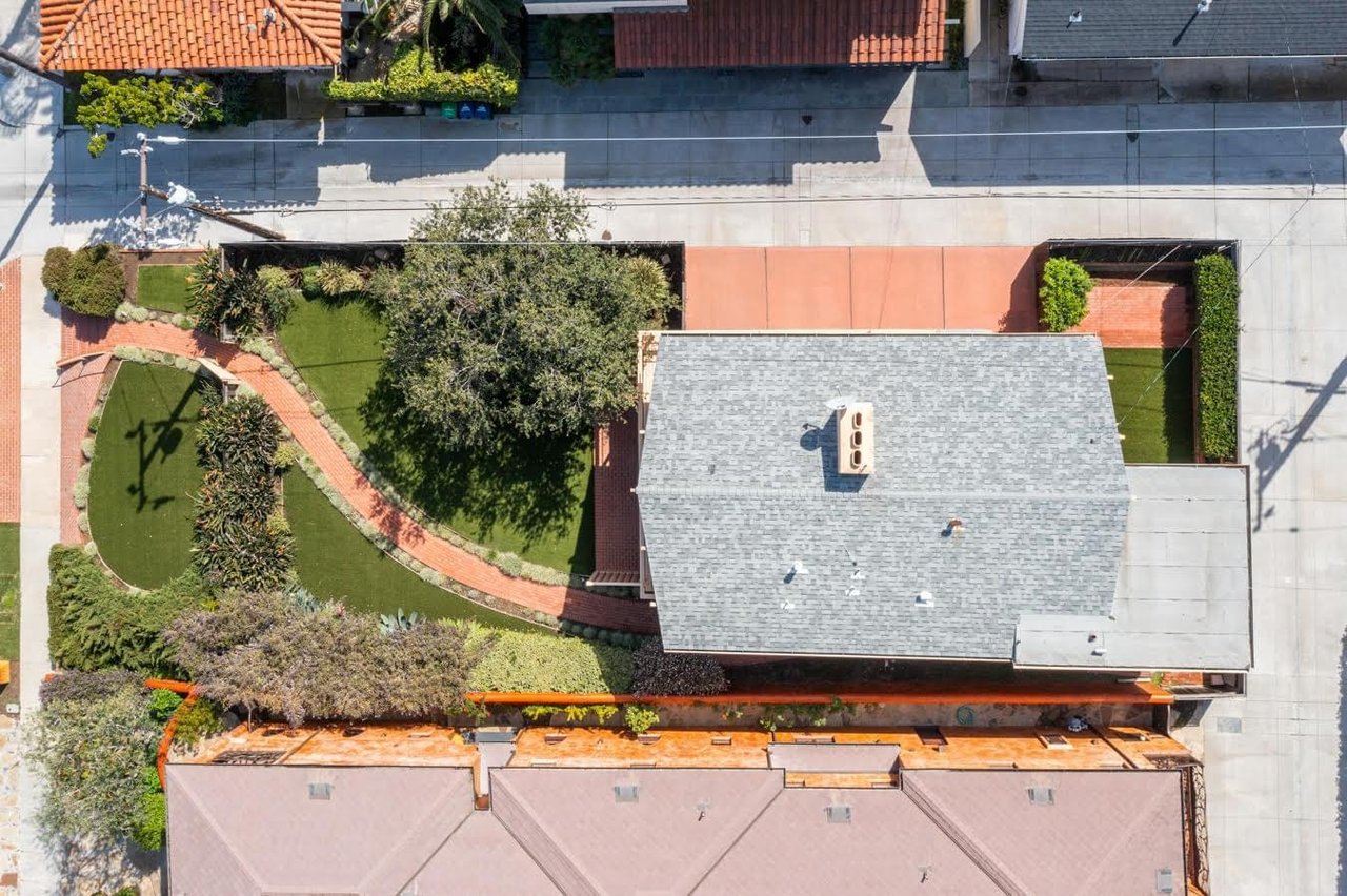  An aerial view of a property with a manicured lawn, a brick pathway, and a gray-roofed house with a chimney.