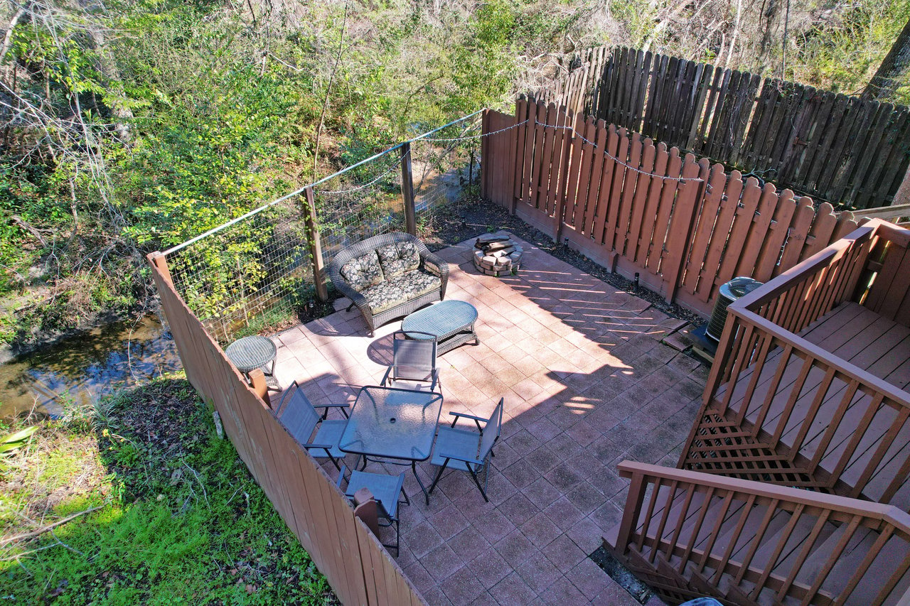 A cozy backyard patio with wicker seating, a small table set, and a fire pit. Surrounded by wooden fencing and lush greenery, sunlit and tranquil.