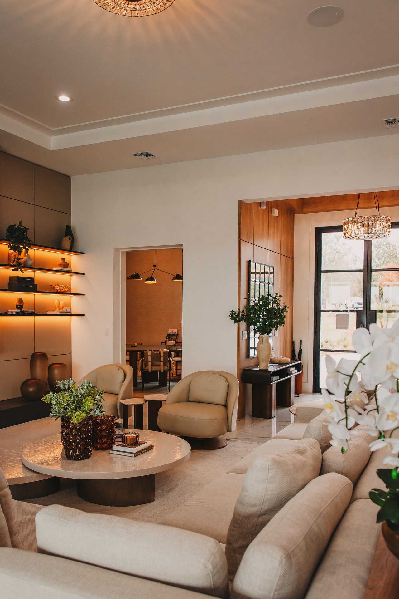 Living room with cream seating, illuminated display shelves, warm wood paneling, and a round coffee table.