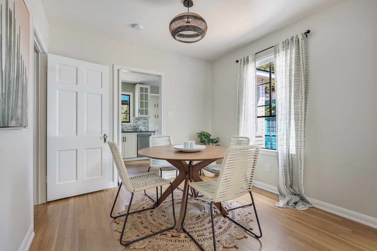Dining room with a round wood table, woven chairs, and a pendant light beside a curtained window.