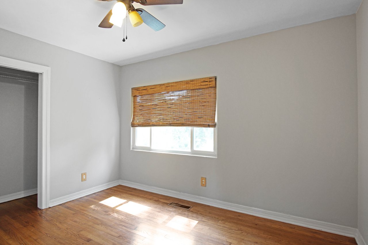 Empty room with light gray walls and wooden floor. A window with bamboo blinds lets in sunlight. Ceiling fan with colorful blades adds a cozy touch.