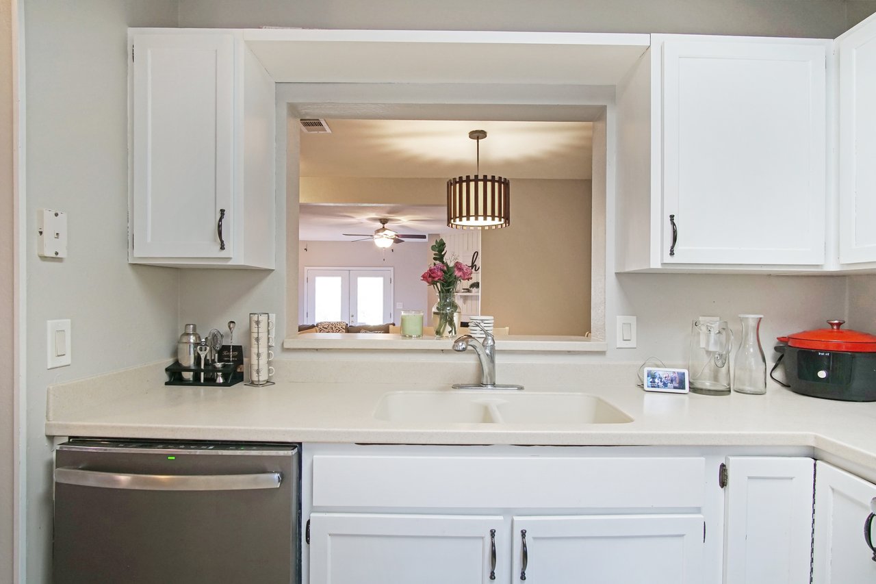 A modern kitchen with white cabinets, a double sink, and a view into a living room. Pink flowers in a vase sit by the sink, creating a cozy ambiance.