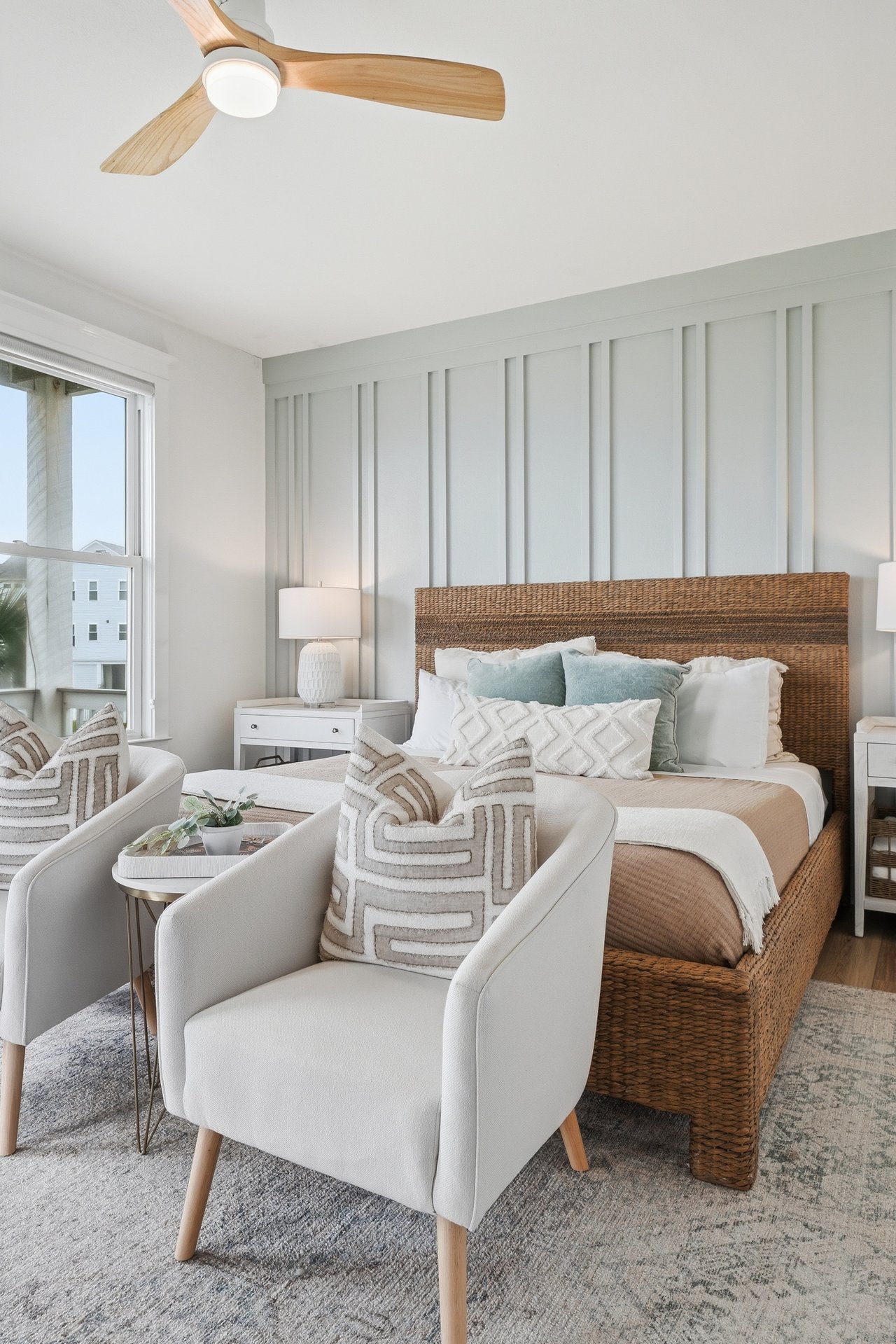 Bright bedroom with a woven rattan bed, pale blue pillows, and upholstered armchairs by a large window.