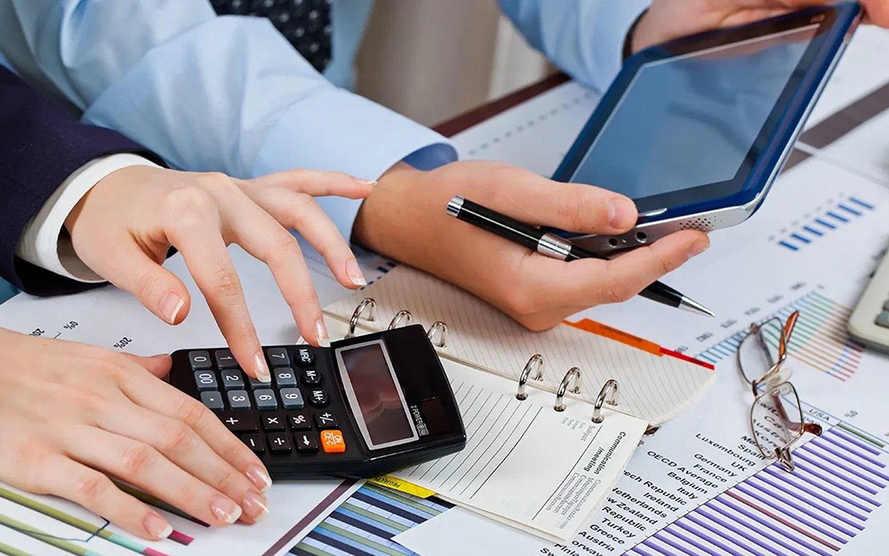 Hands use a calculator and tablet while reviewing financial charts and paperwork.