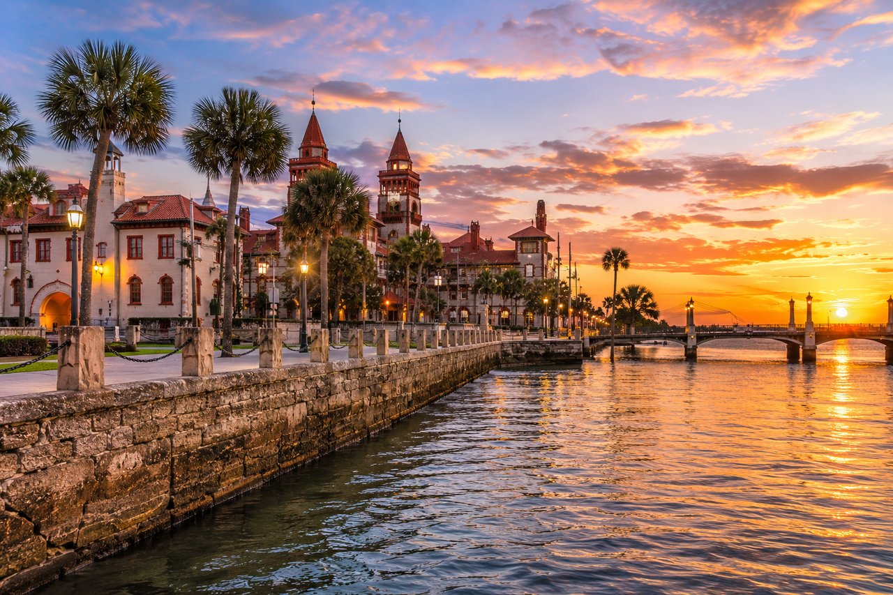 Historic waterfront in St. Augustine, Florida, featuring Spanish colonial architecture, palm trees, scenic bayfront, stone seawall, and a colorful sunset over the water.