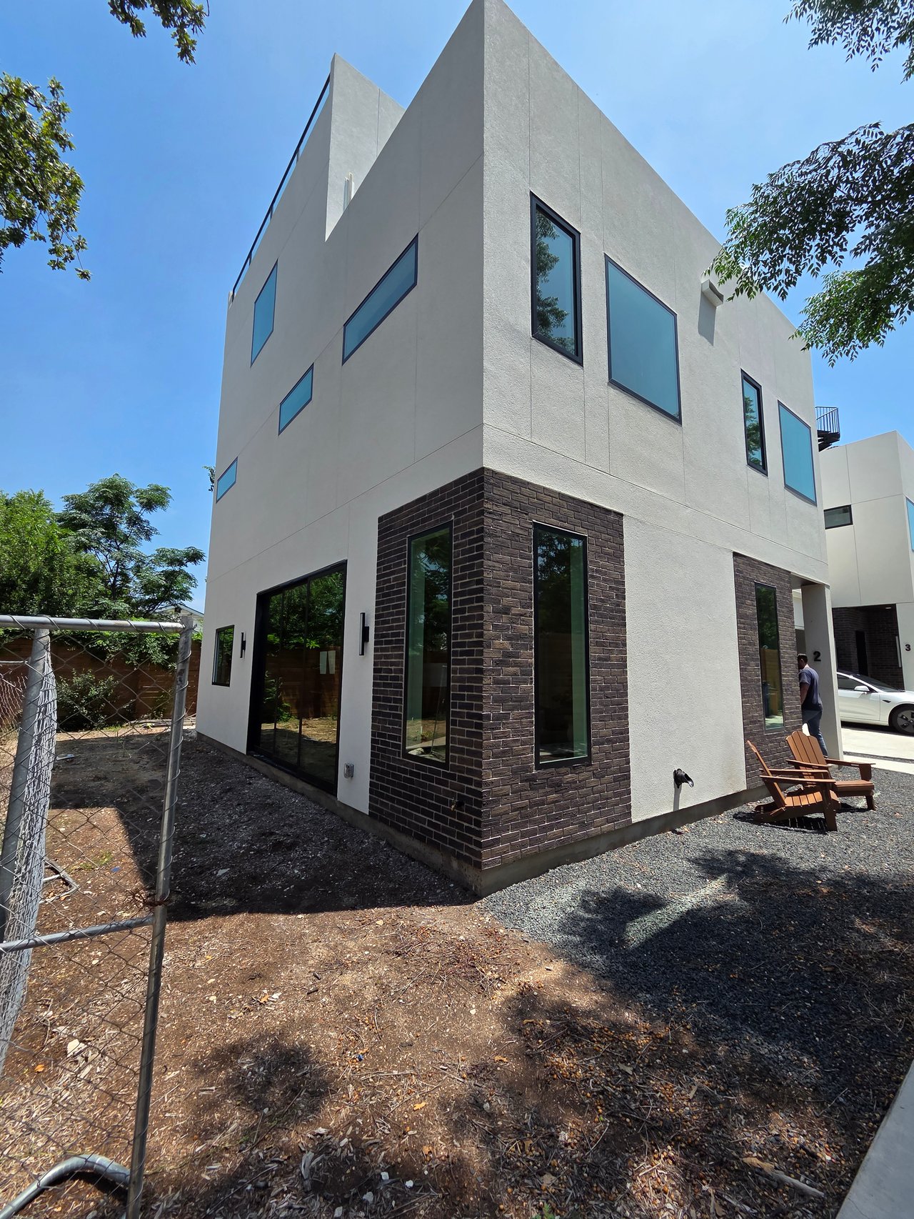 Modern townhouse with white stucco, dark brick accents, and tall angular windows beside a gravel yard.