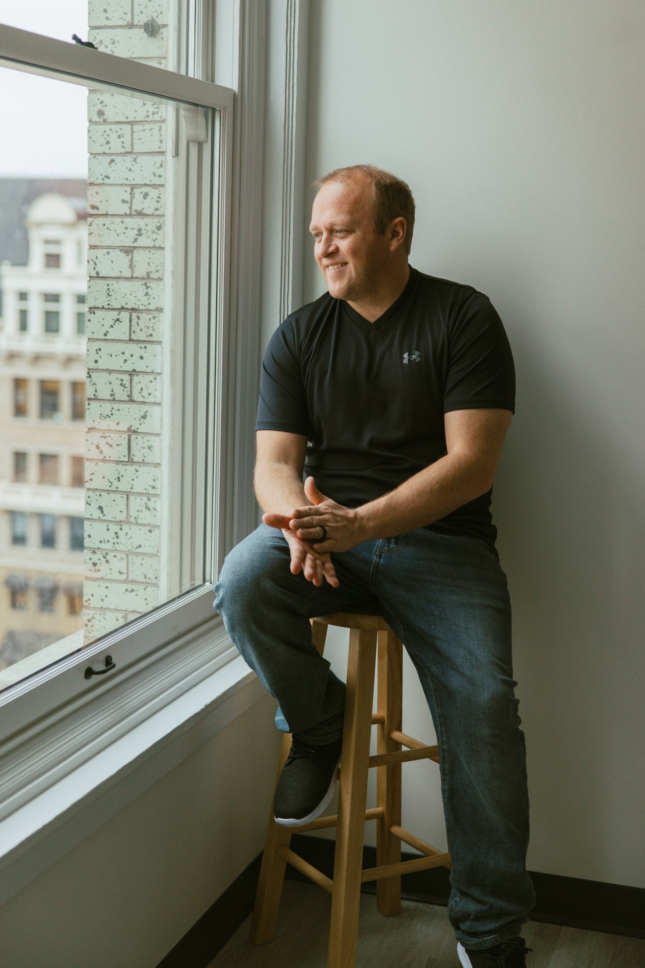 Mike Helmberger seated on a wooden stool beside a large window.