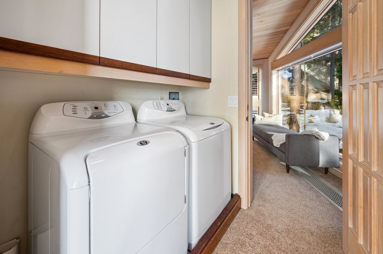 Laundry room with white washer and dryer beside a windowed sitting room overlooking snow and trees.