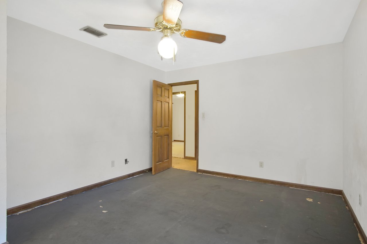 Empty room with white walls and dark floor, featuring a wooden door slightly ajar. A ceiling fan with a light illuminates the space.