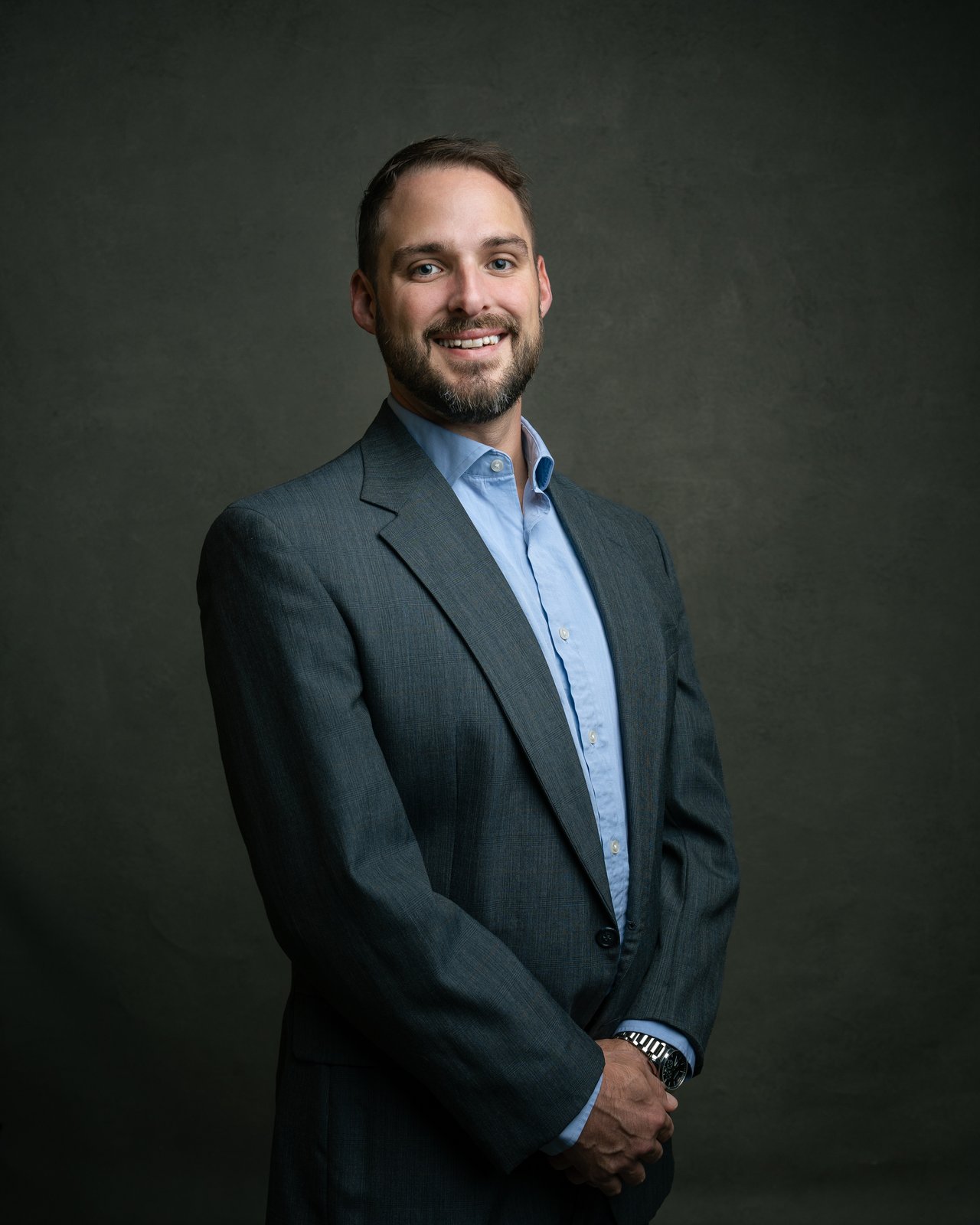 Jacob K. Sheets in a charcoal suit and light blue shirt, smiling with his hands clasped.