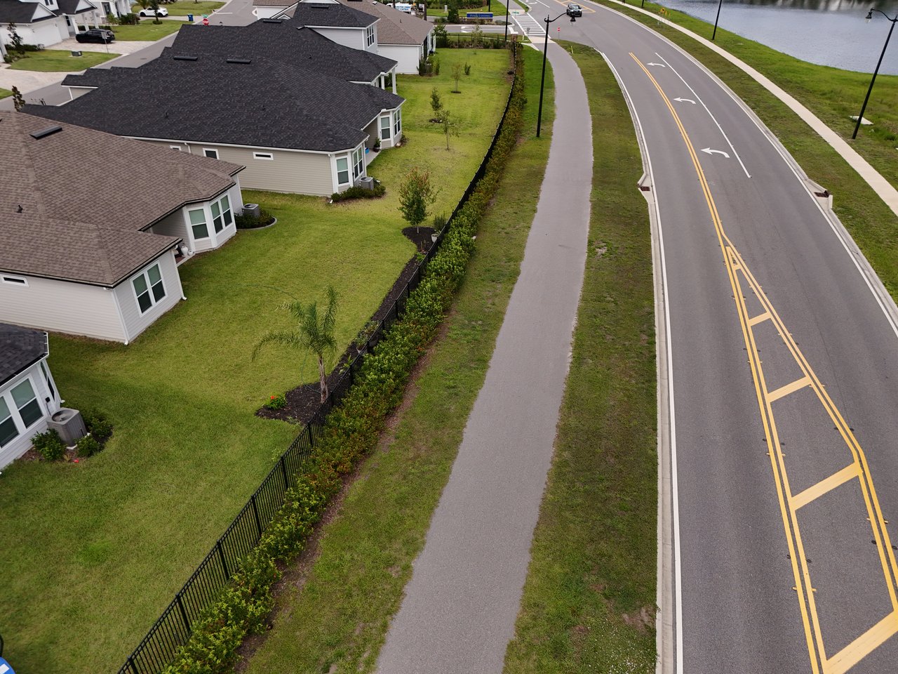 Aerial view of neighborhood homes beside a fenced lawn, sidewalk, and curving road.