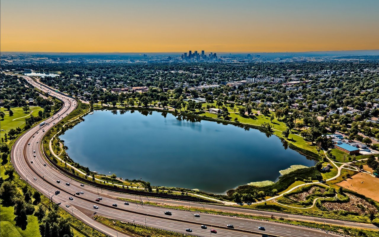 Aerial view of a lake, parkland, and curving freeway with a distant downtown skyline.
