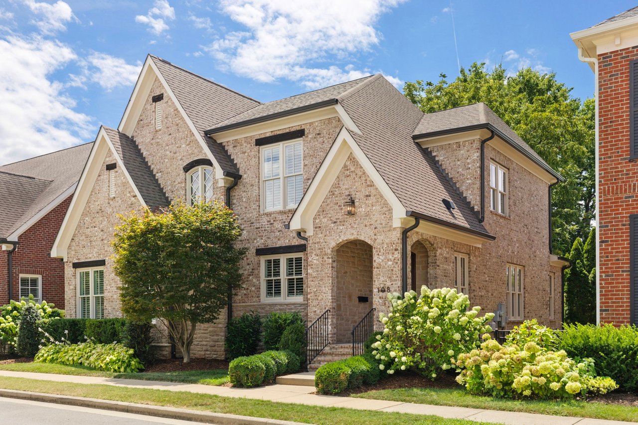 Two-story brick house with steep gables, an arched entry, and flowering shrubs along the front.