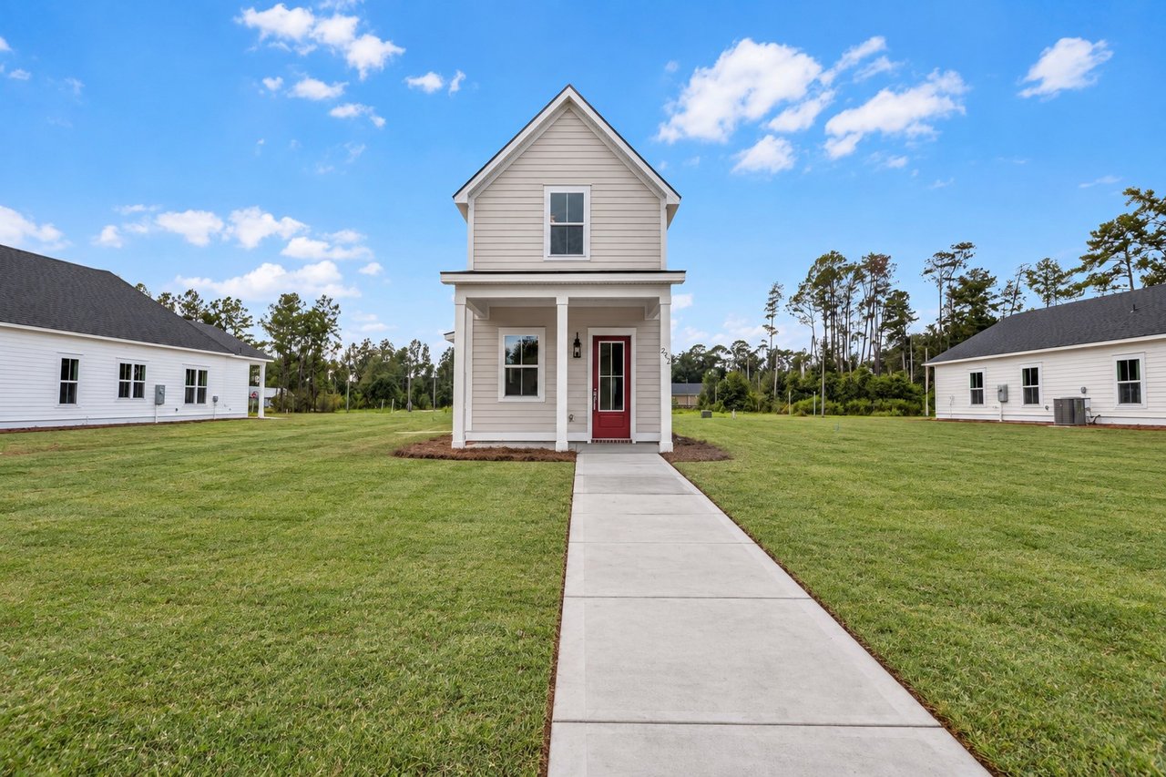 Two-story beige home with a red front door and concrete walkway across a broad lawn.
